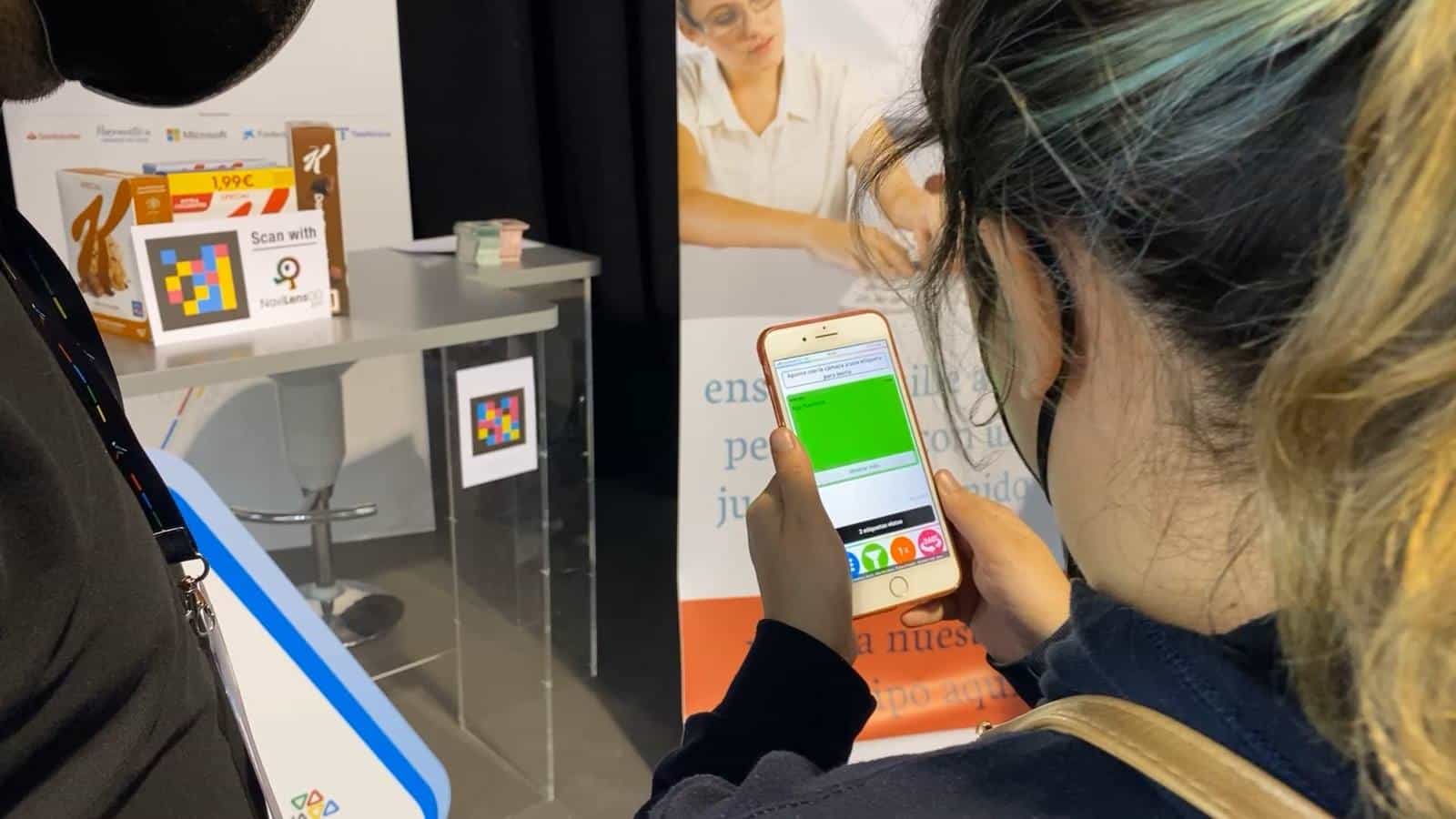 A Tifloinnova visitor scans with her iPhone a NaviLens code placed on a plinth at stand 12; next to the code, a “Scan with NaviLens GO” sign and two Special K Classic cereal boxes with a NaviLens code and a 1.99 € price tag. The phone screen shows the scan result on a green strip with the text “Stop NaviLens”