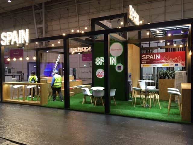 Vista general del Pabellón de España en el Web Summit 2021 de Lisboa: estructura modular de madera y acero negro con letras luminosas «SPAIN», césped artificial verde, mesas y sillas blancas, bombillas tipo Edison y, al fondo, los logos institucionales de Gobierno de España, Red.es e ICEX