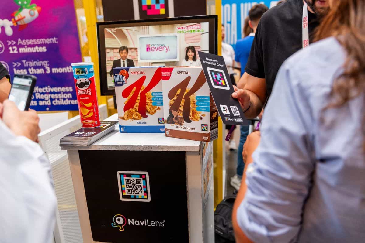 Stand de NaviLens en el South Summit Madrid 2024: sobre un mostrador blanco y negro con el logotipo de NaviLens, dos cajas de cereales Special K —Classic, en azul, y Dark Chocolate, en marrón— junto a un bote rojo de Pringles Original, todos con códigos NaviLens visibles; detrás, una pantalla emite un informativo de TV japonés mostrando códigos NaviLens; en primer plano, una visitante sostiene una tarjeta «NaviLens Accessible Code» con enlaces a Google Play y App Store, y otro visitante escanea con su móvil el código pegado en la peana del stand