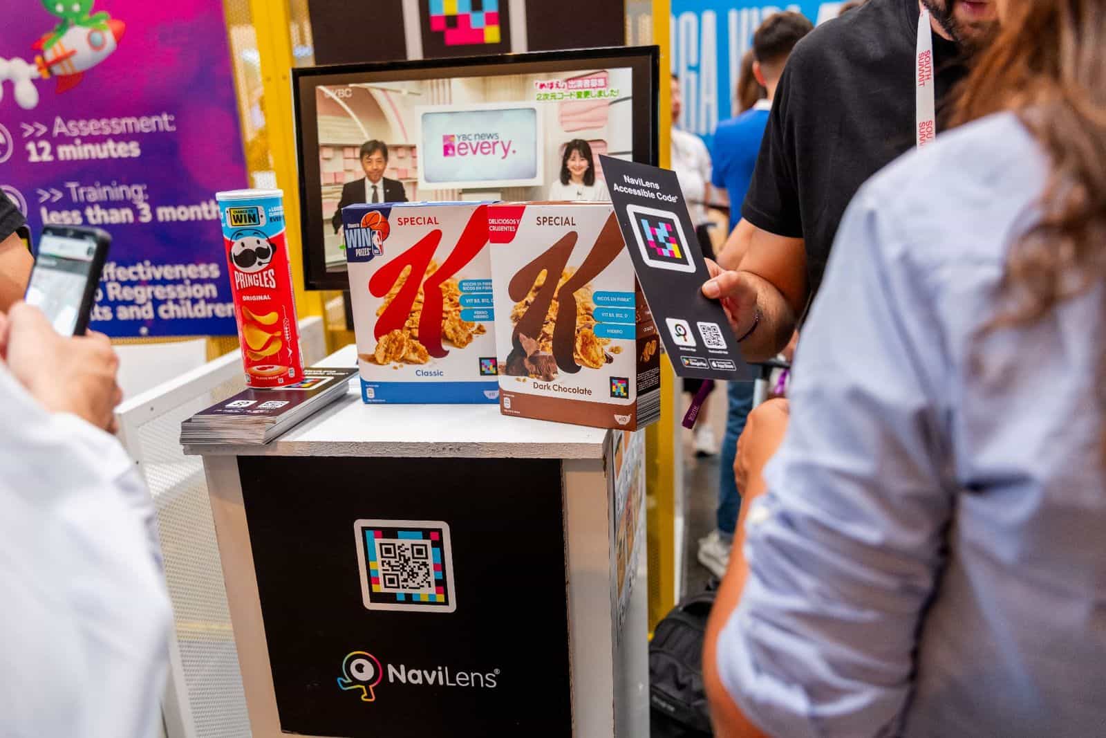 Stand di NaviLens al South Summit Madrid 2024: su un bancone bianco e nero con il logo di NaviLens, due confezioni di cereali Special K —Classic, in azzurro, e Dark Chocolate, in marrone— accanto a un tubo rosso di Pringles Original, tutti con codici NaviLens visibili; dietro, uno schermo trasmette un telegiornale giapponese che mostra codici NaviLens; in primo piano una visitatrice tiene una card «NaviLens Accessible Code» con i link a Google Play e App Store, e un altro visitatore inquadra con il telefono il codice applicato sul basamento dello stand