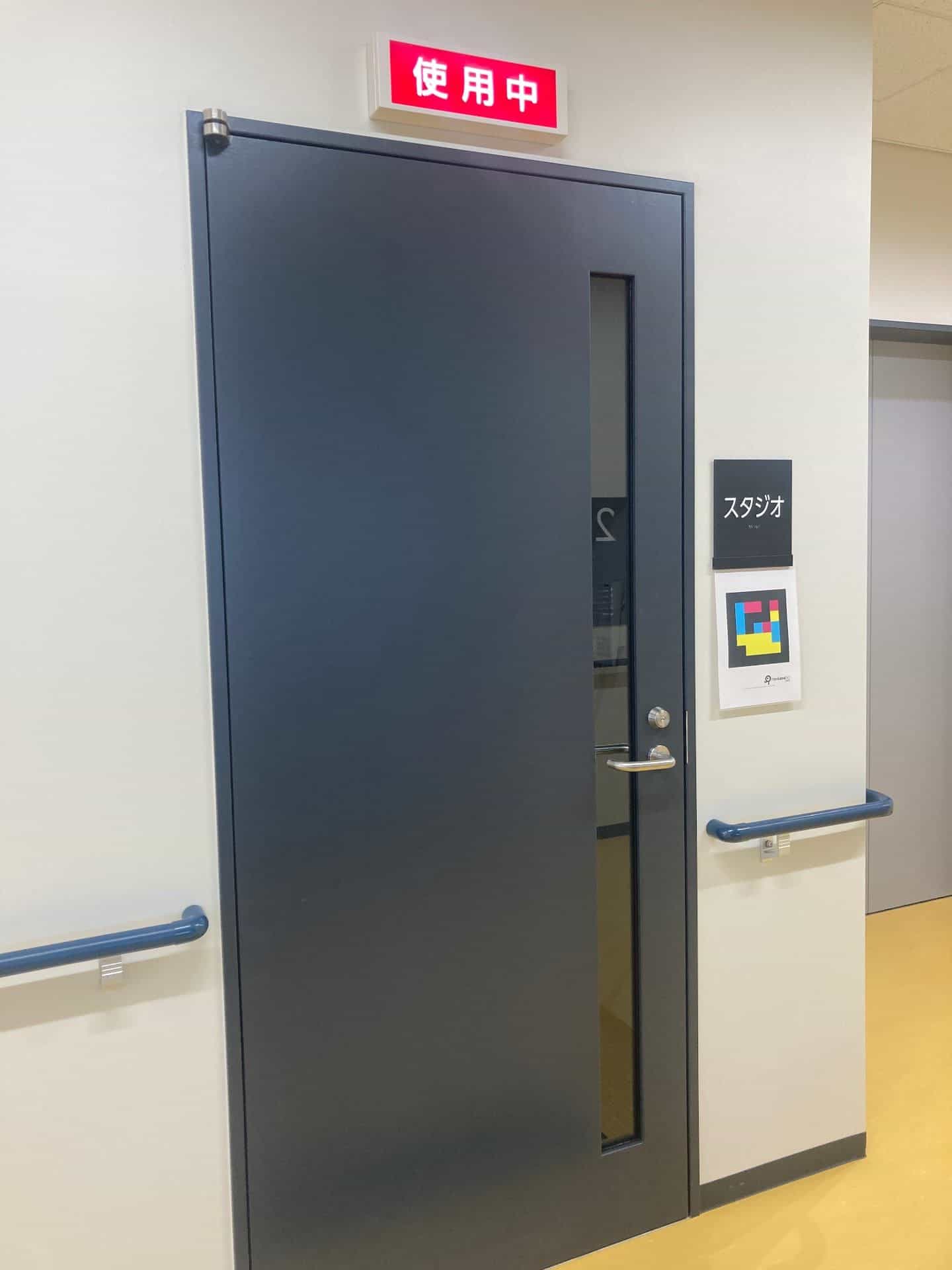 Porta a battente blu scuro quasi nera dello studio di registrazione («スタジオ»): maniglione metallico verticale con serratura, spioncino rettangolare verticale e, sopra l’architrave, un’insegna rossa illuminata «使用中» (in uso); a destra della porta, targa tattile nera con il testo bianco «スタジオ» e, subito sotto, un cartello A4 con il codice NaviLens GO in ciano, magenta e giallo su fondo nero; corrimano blu su entrambi i lati del corridoio in vinile giallo