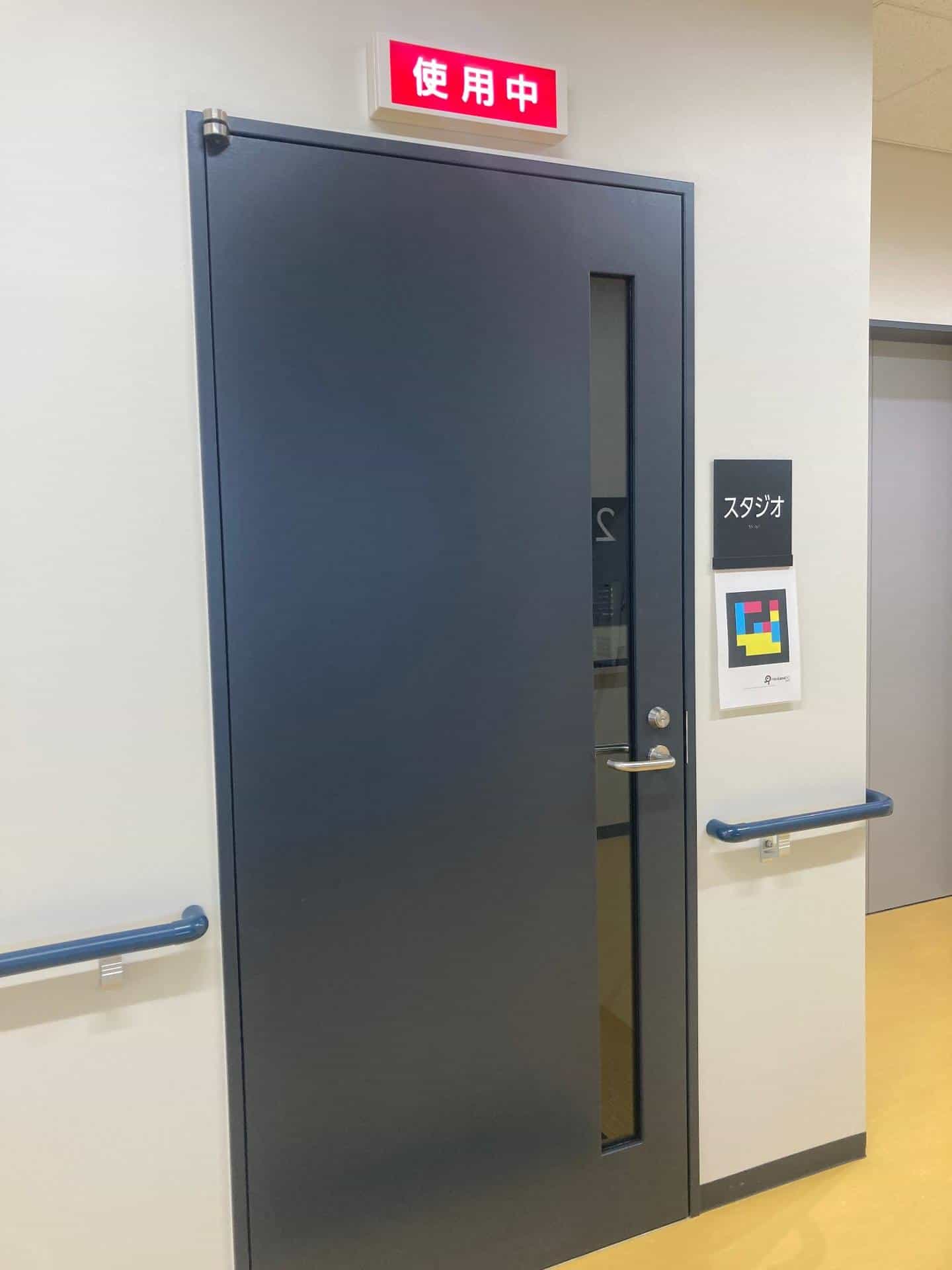 Almost-black dark-blue swing door of the recording studio (“スタジオ”): vertical metal pull handle with lock, vertical rectangular peephole and, above the lintel, a red illuminated “使用中” (in use) sign; to the right of the door, a black tactile plate with white “スタジオ” text and, just below, an A4 sign with the NaviLens GO code in cyan, magenta and yellow on black; blue handrails on both sides of the yellow vinyl corridor