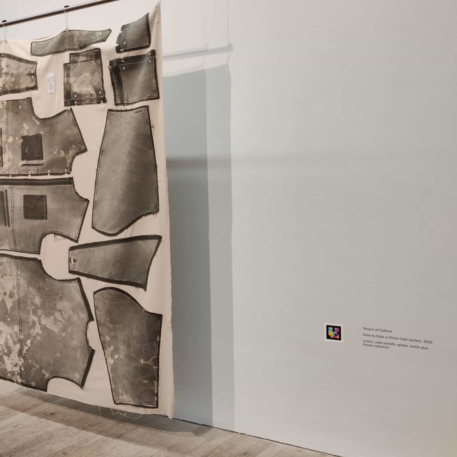 Textile piece How to Style a Chore Coat (series), 2020 by Tenant of Culture, hung in a room, with its caption and a NaviLens code on the adjacent wall