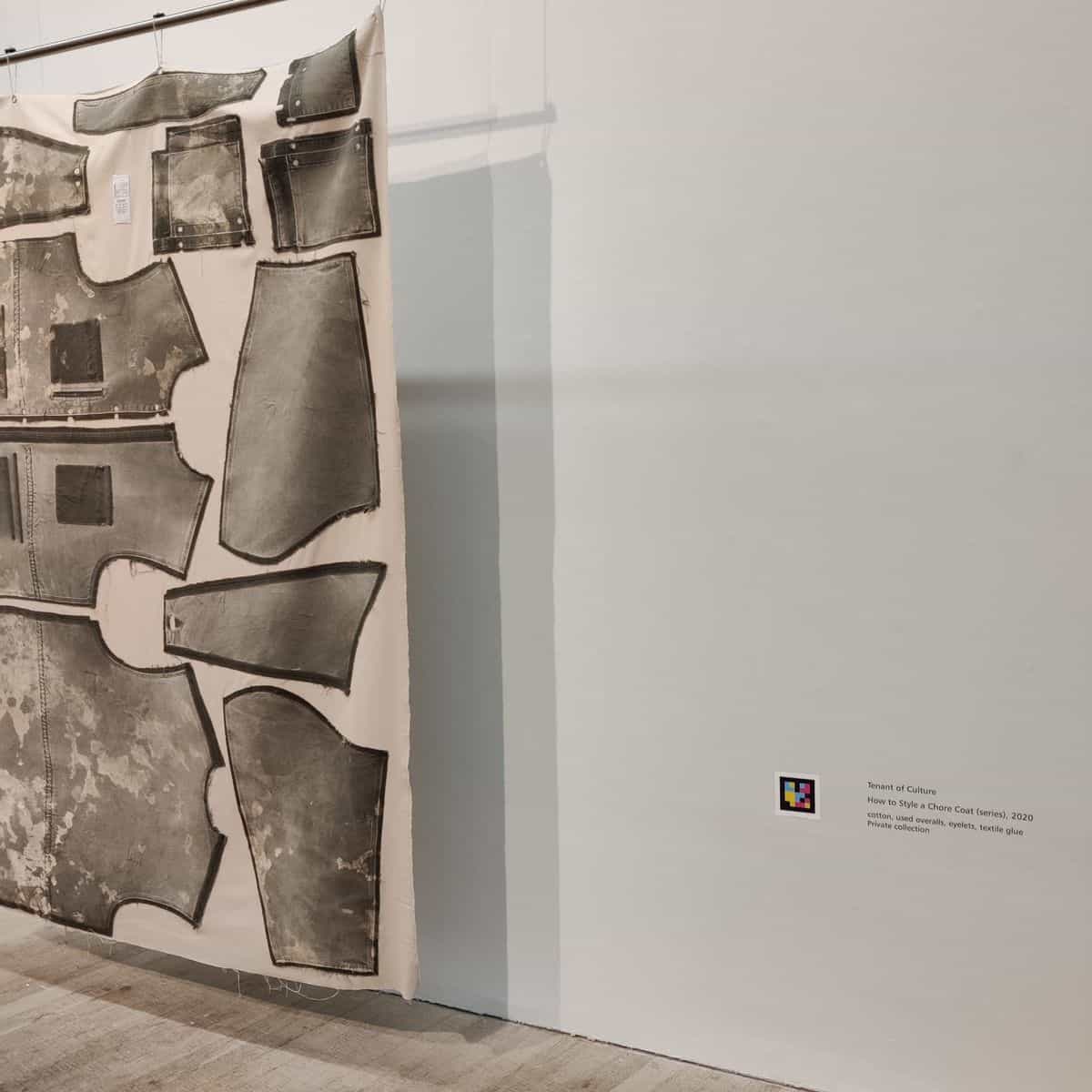 Textiles Kunstwerk „How to Style a Chore Coat (series), 2020“ von Tenant of Culture, in einem Raum hängend, mit Bildunterschrift und einem NaviLens-Code an der angrenzenden Wand