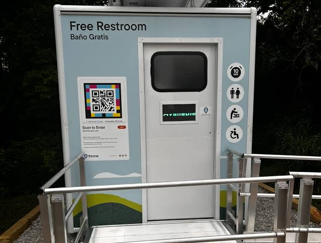 Vista frontal de una cabina Throne con el rótulo «Free Restroom · Baño Gratis», pictogramas de aseo accesible, cambiador y aforo, y un cartel «Scan to Enter» con un código NaviLens a la izquierda de la puerta.