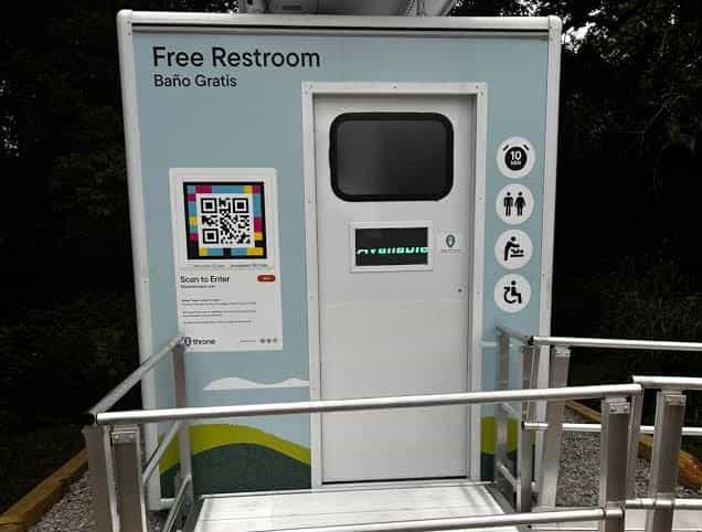 Front view of a Throne unit with a «Free Restroom · Baño Gratis» sign, accessible-toilet, changing-table and occupancy pictograms, and a «Scan to Enter» sign with a NaviLens code to the left of the door.