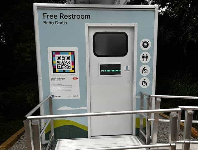 Front view of a Throne unit with a “Free Restroom · Baño Gratis” sign, accessible-toilet, changing-table and occupancy pictograms, and a “Scan to Enter” sign with a NaviLens code to the left of the door.