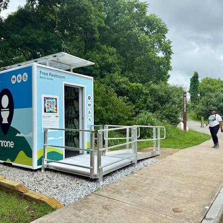 Cabina Throne con la puerta abierta mostrando el interior, vista lateral con el rótulo «Free Restroom · Baño Gratis», ilustración de un inodoro y un código NaviLens junto a la entrada.