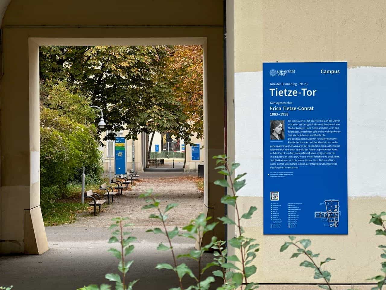 Tafel „Tore der Erinnerung – Nr. 23 Tietze-Tor“ für die Kunsthistorikerin Erica Tietze-Conrat (1883–1958), rechts neben einem Torbogen zum Campushof mit Holzbänken und grünen Bäumen