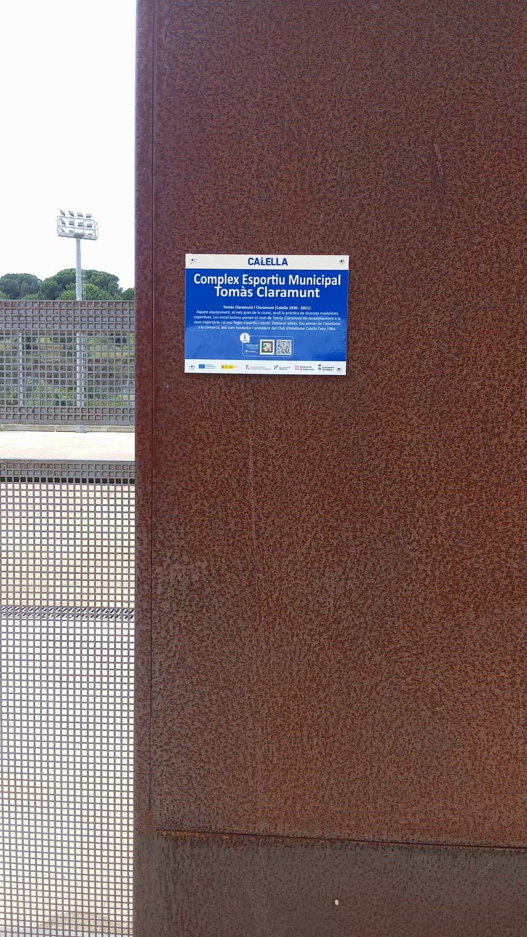 Placa azul CALELLA del Complex Esportiu Municipal Tomàs Claramunt con código NaviLens sobre panel de acero corten