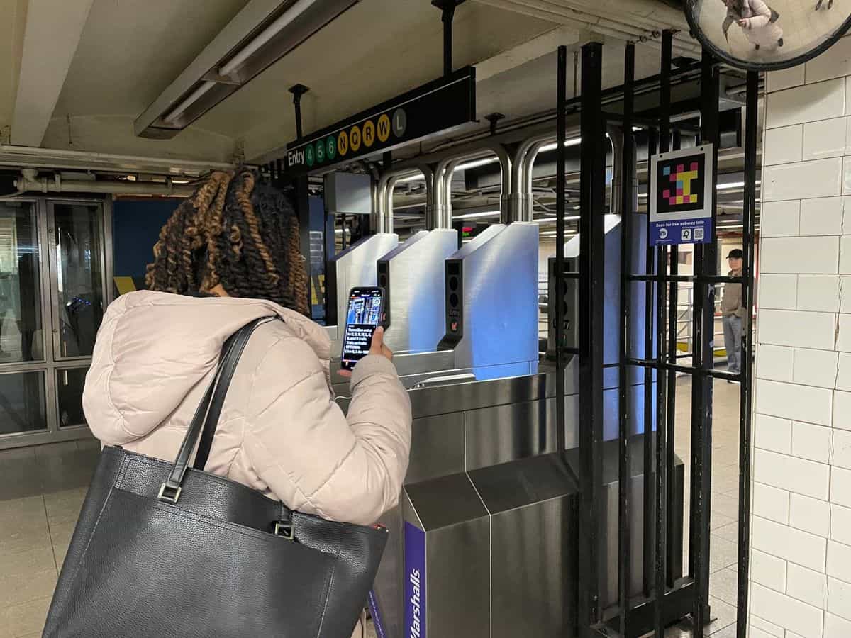 MTA turnstile bank with overhead sign «Entry 4 5 6 N Q R W L», NaviLens code high on the right and a person scanning with their phone; in the background, the 14 St-Union Square concourse