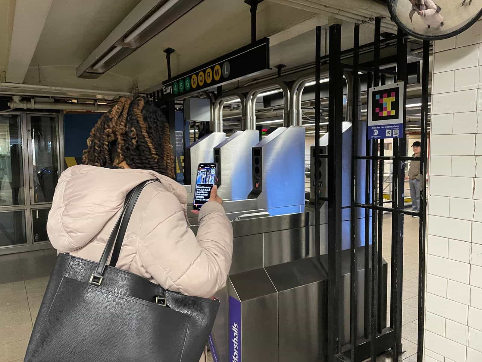 MTA turnstile bank with overhead sign “Entry 4 5 6 N Q R W L”, NaviLens code high on the right and a person scanning with their phone; in the background, the 14 St-Union Square concourse