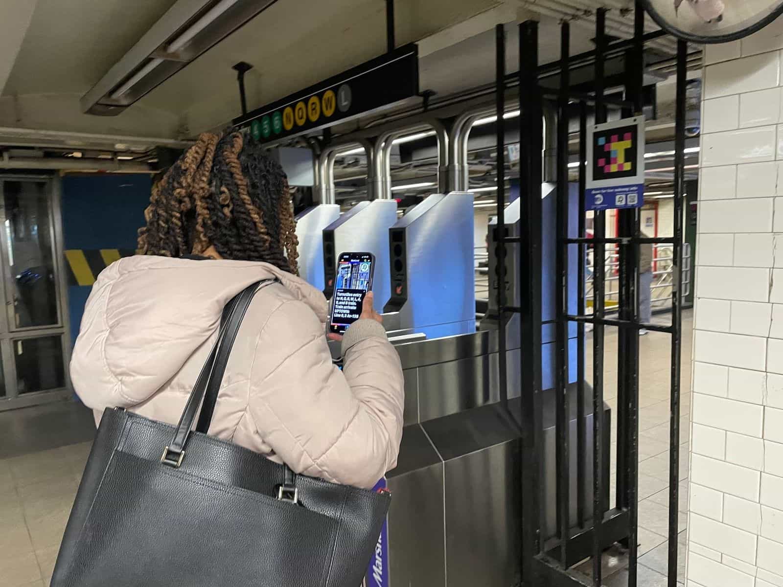 Person walks up with the phone to the NaviLens code mounted at eye level on the metal partition, next to the turnstiles labeled “N Q R W L” at 14 St-Union Square