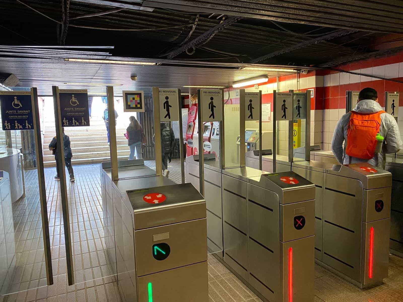 Linea dei tornelli «Pas individual» dell’atrio con pannelli azzurri «Junts davant» a sinistra e, sul muro dietro, un codice NaviLens quadrato a colori che informa sui passaggi prioritari per sedie a rotelle, bambini o valigie e sull’ingresso in banchina