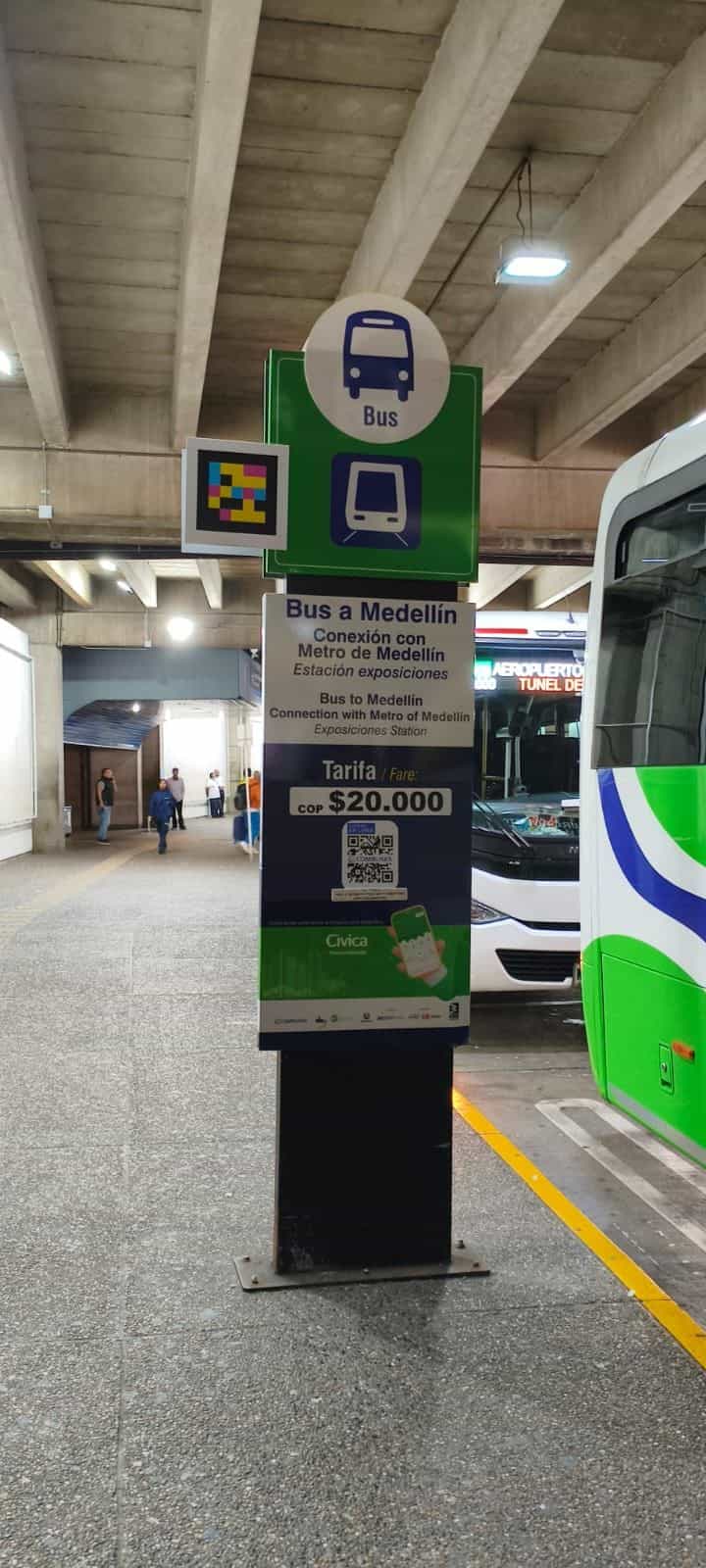 Totem «Bus a Medellín» im Bodentransportbereich des Flughafens José María Córdova: grünes Schild mit runden Bus- und Metro-Piktogrammen und links davon quadratischer NaviLens-Code in Cyan, Magenta und Gelb; darunter Tafel «Bus nach Medellín · Anschluss an die Metro Medellín · Station Exposiciones» mit Tarif «COP 20.000» und COMBUSES-QR-Code; neben dem Totem ein grün-weißer Bus der Linie «AEROPUERTO TÚNEL DE…»