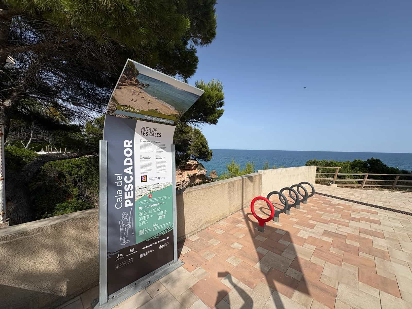 Geschwungenes Totem der Ruta de les Cales an der Cala del Pescador: Foto der Bucht im oberen Abschluss, Pfeile zur Cala Bot und zur Cala dels Àngels, dreisprachiger Text und ein NaviLens-Code, auf der gepflasterten Promenade mit dem Meer im Hintergrund