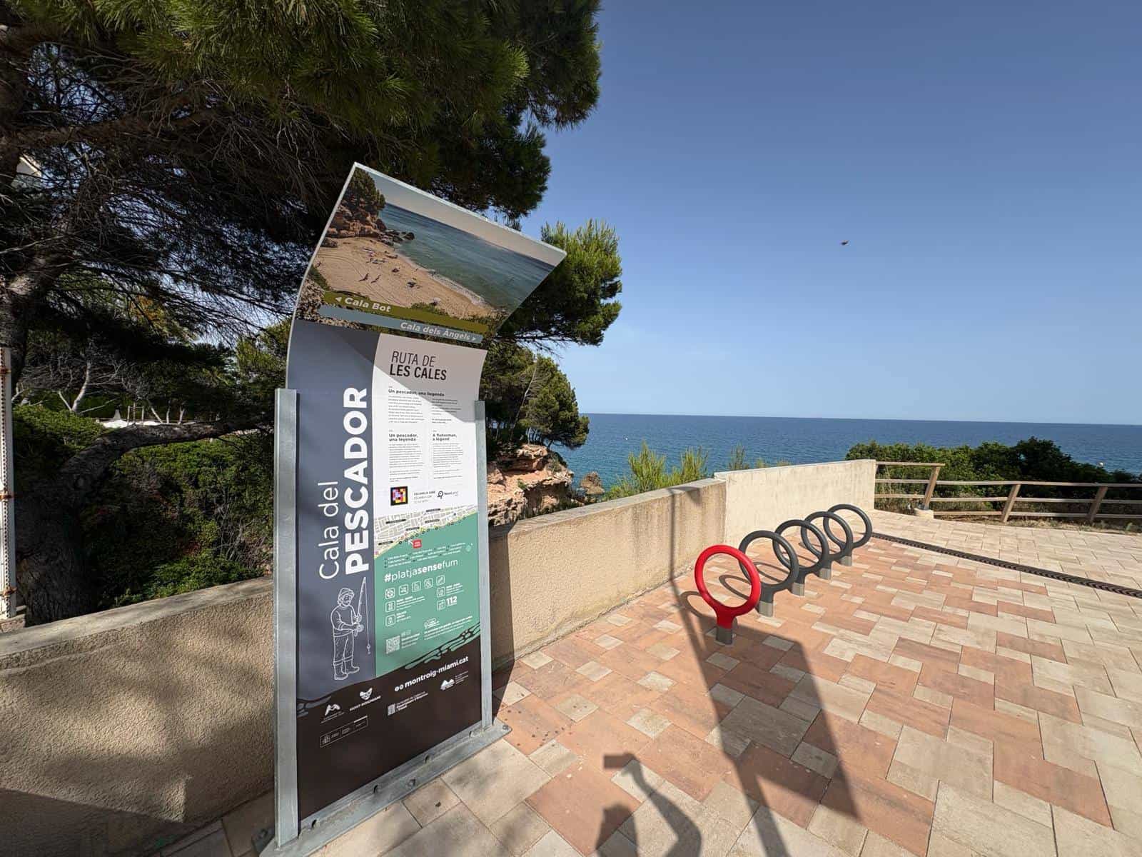 Curved Ruta de les Cales totem at Cala del Pescador: a photo of the cove along the top, arrows towards Cala Bot and Cala dels Àngels, trilingual text and a NaviLens code, on the paved promenade with the sea behind