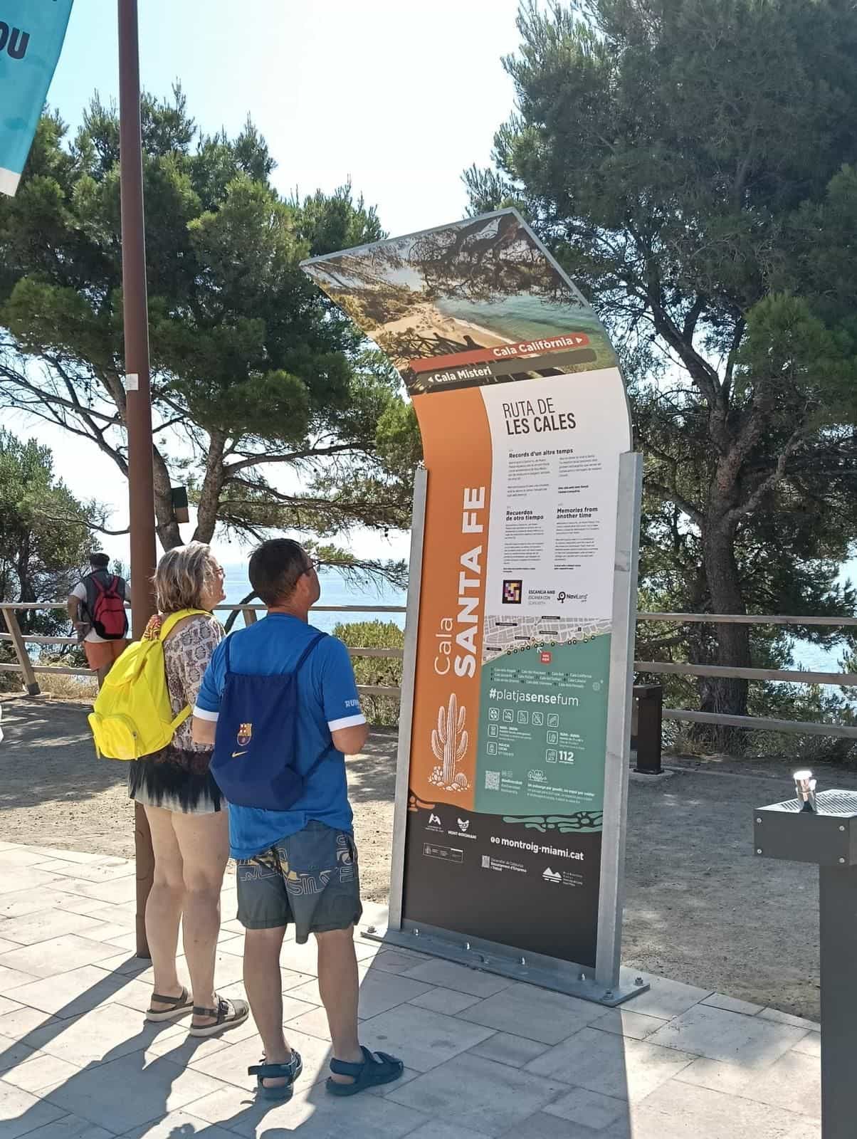 Zwei Besucher mit Rucksäcken vor dem Totem der Cala Santa Fe mit der Geschichte «Records d'un altre temps» und dem NaviLens-Code