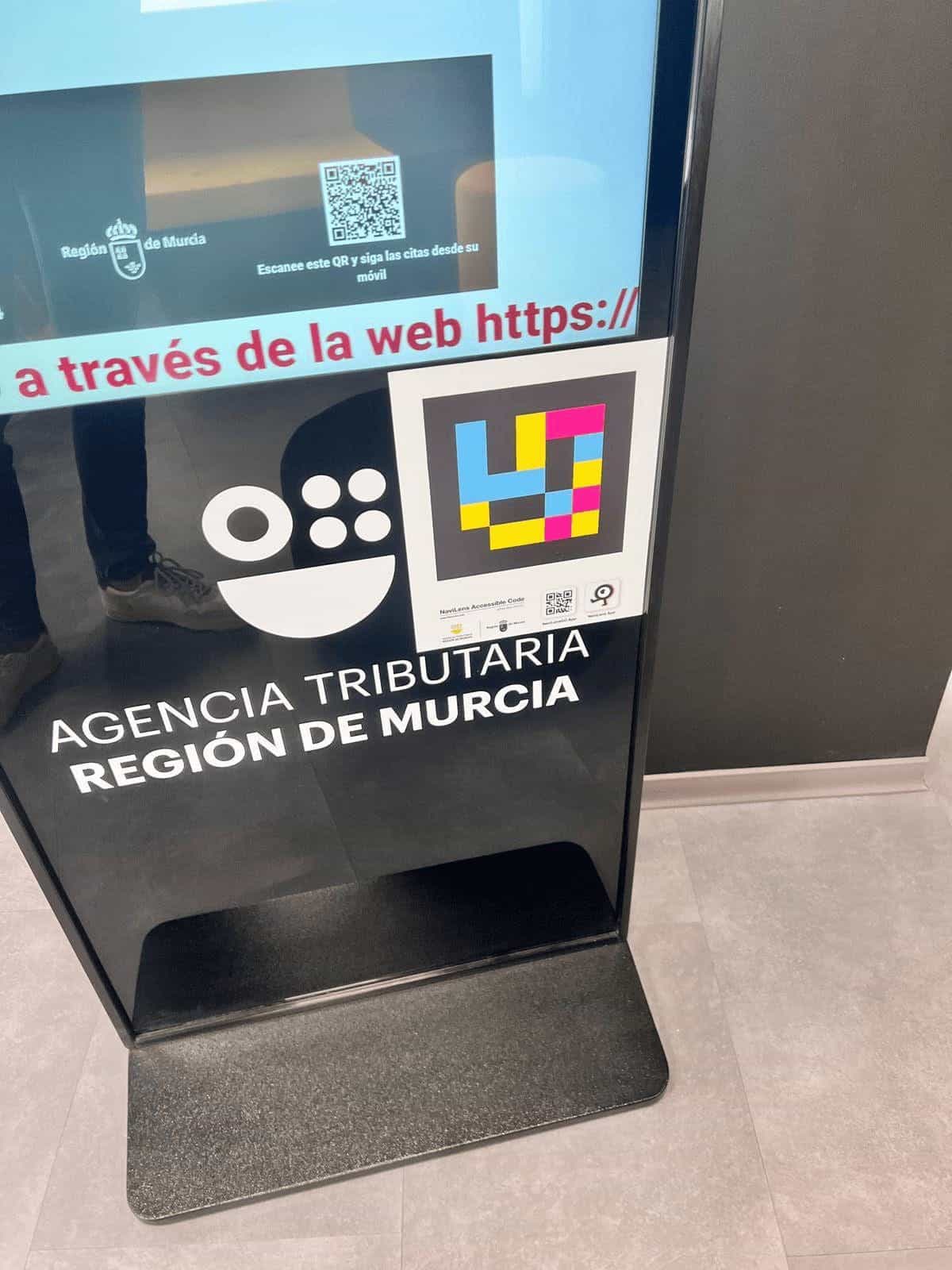 Tótem digital de gestión de citas con pantalla azul, código QR para seguir las citas desde el móvil y código NaviLens cuadrado en cian, magenta y amarillo en el frontal.