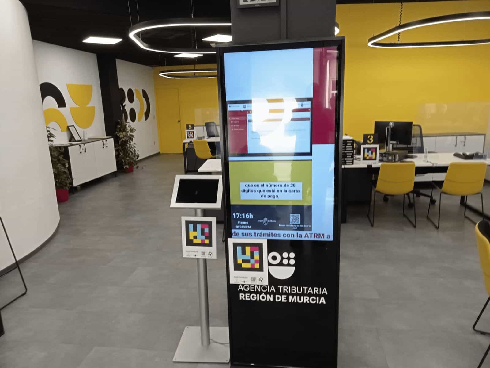 Atrio dell’ufficio ATRM di Águilas nel suo modello «store»: totem digitale nero verticale con schermo acceso in blu, granata e verde che mostra l’avviso «que es el número de 28 dígitos que está en la carta de pago» e l’ora «17:16h Viernes 28/06/2024»; sotto il totem, sul frontale nero, il codice NaviLens quadrato in ciano, magenta e giallo e i logo «AGENCIA TRIBUTARIA · REGIÓN DE MURCIA».