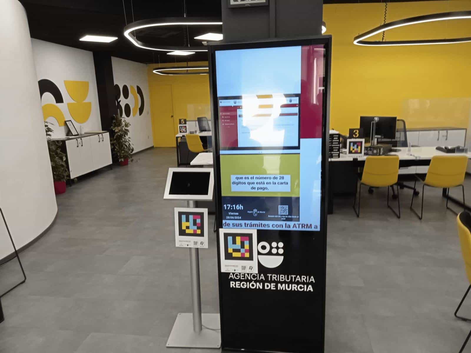 Lobby of the ATRM Águilas “store” office: vertical black ticket totem with screen on, NaviLens code on its black front and ATRM · Region of Murcia logos.