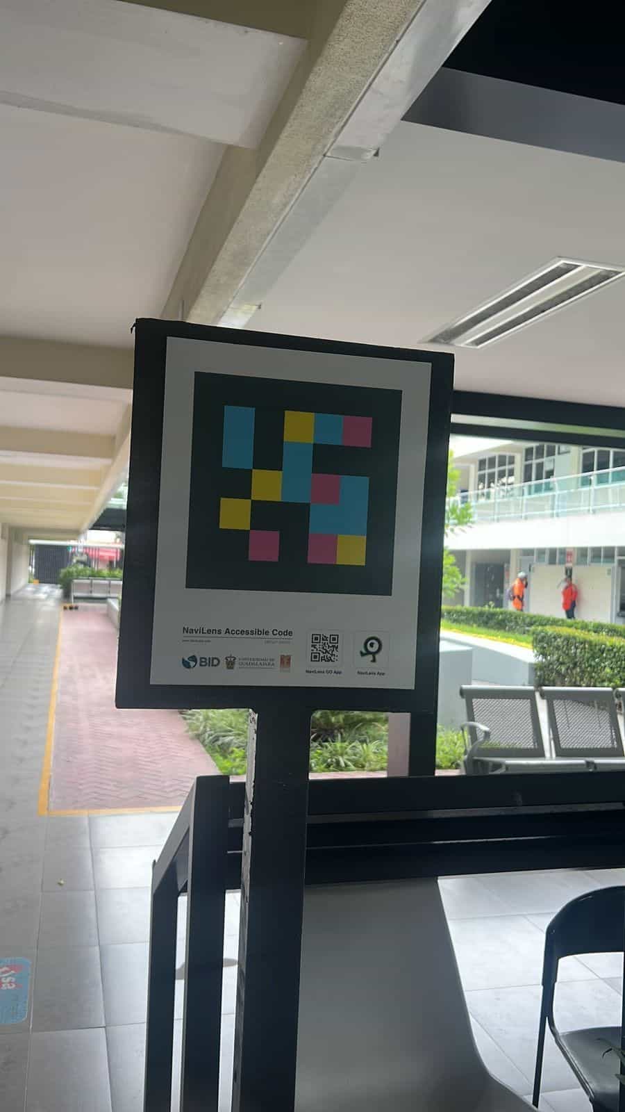 Vertical totem with a large NaviLens code and the IDB, UdeG and CUCS logos installed in an open campus corridor.