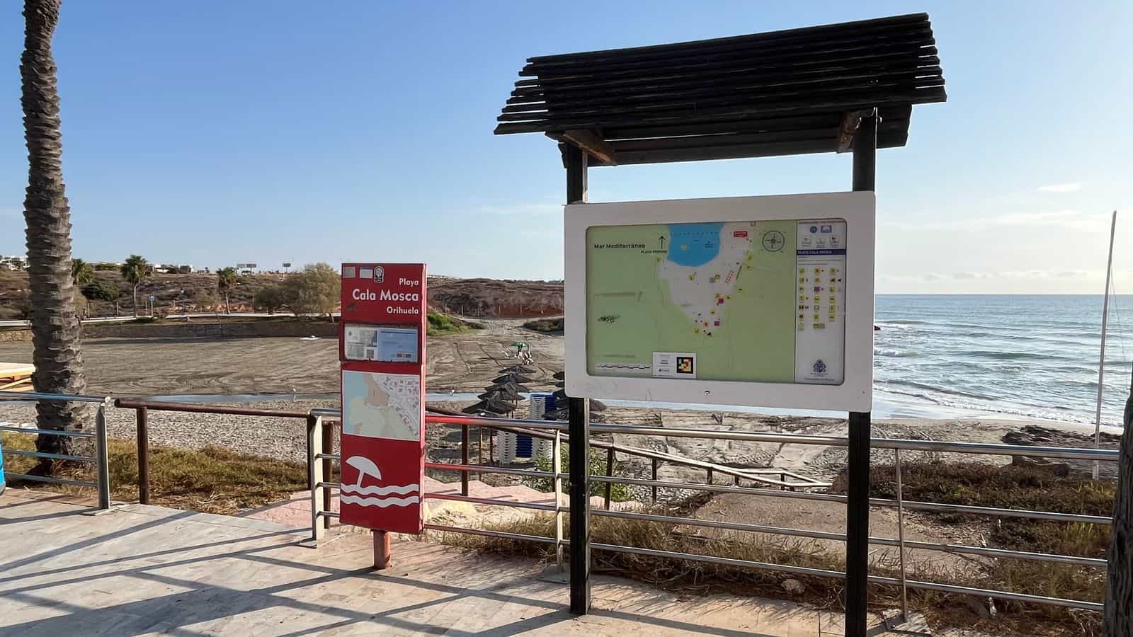 Aussichtspunkt am Strand Cala Mosca in Orihuela: links das rote Totem „Playa Cala Mosca · Orihuela“ mit den Gütesiegeln des Tourismus; rechts, unter einer Pergola aus Holzlatten, die weiße Tafel mit dem Lageplan der Bucht und dem Schild mit dem NaviLens-Code, dahinter das Mittelmeer
