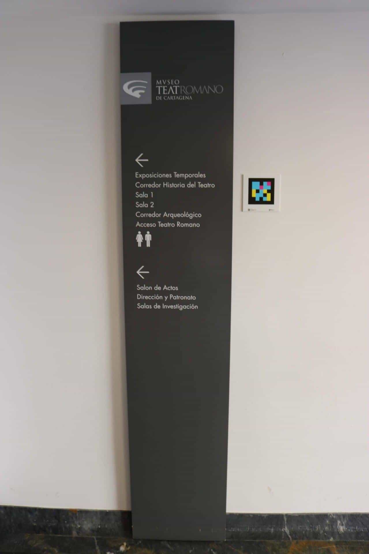 Black directional totem of MUSTER showing routes to Temporary Exhibitions, History of the Theatre Corridor, Rooms 1 and 2, Archaeological Corridor, Access to the Roman Theatre and Assembly Hall, with a NaviLens code beside it
