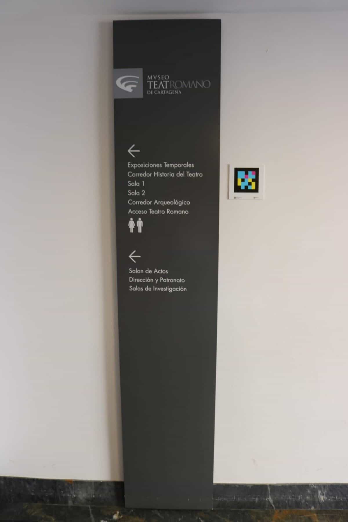 Schwarze Wegweiser-Stele des MUSTER mit Hinweisen auf Sonderausstellungen, Korridor Geschichte des Theaters, Säle 1 und 2, Archäologischer Korridor, Zugang zum Römischen Theater und Veranstaltungssaal; daneben ein NaviLens-Code
