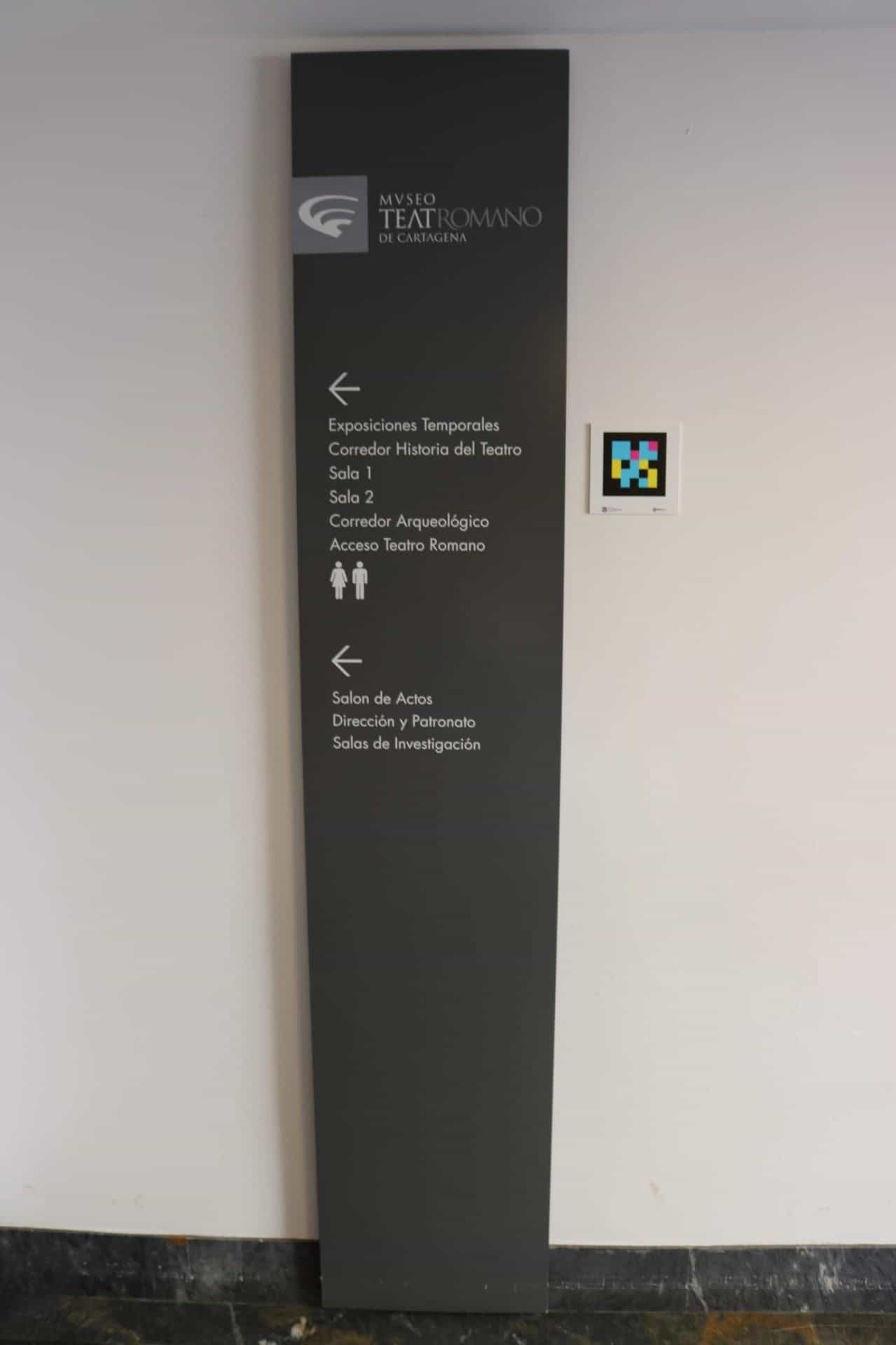 Schwarze Wegweiser-Stele des MUSTER mit Hinweisen auf Sonderausstellungen, Korridor Geschichte des Theaters, Säle 1 und 2, Archäologischer Korridor, Zugang zum Römischen Theater und Veranstaltungssaal; daneben ein NaviLens-Code