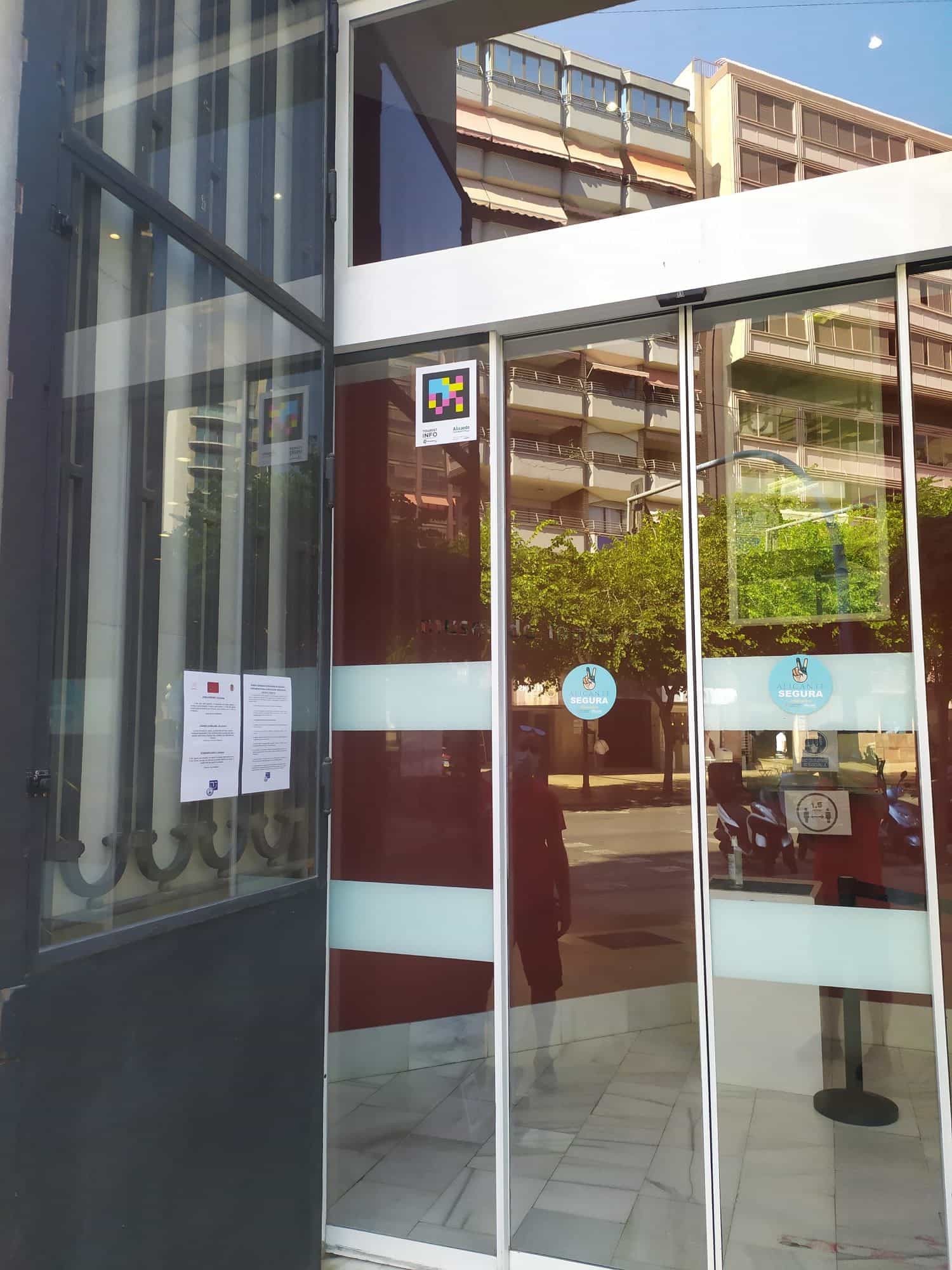 Verglaster Eingang des Tourist-Info-Büros in Alicante mit NaviLens-Code an der Tür und COVID-Aushang „Alicante Segura”