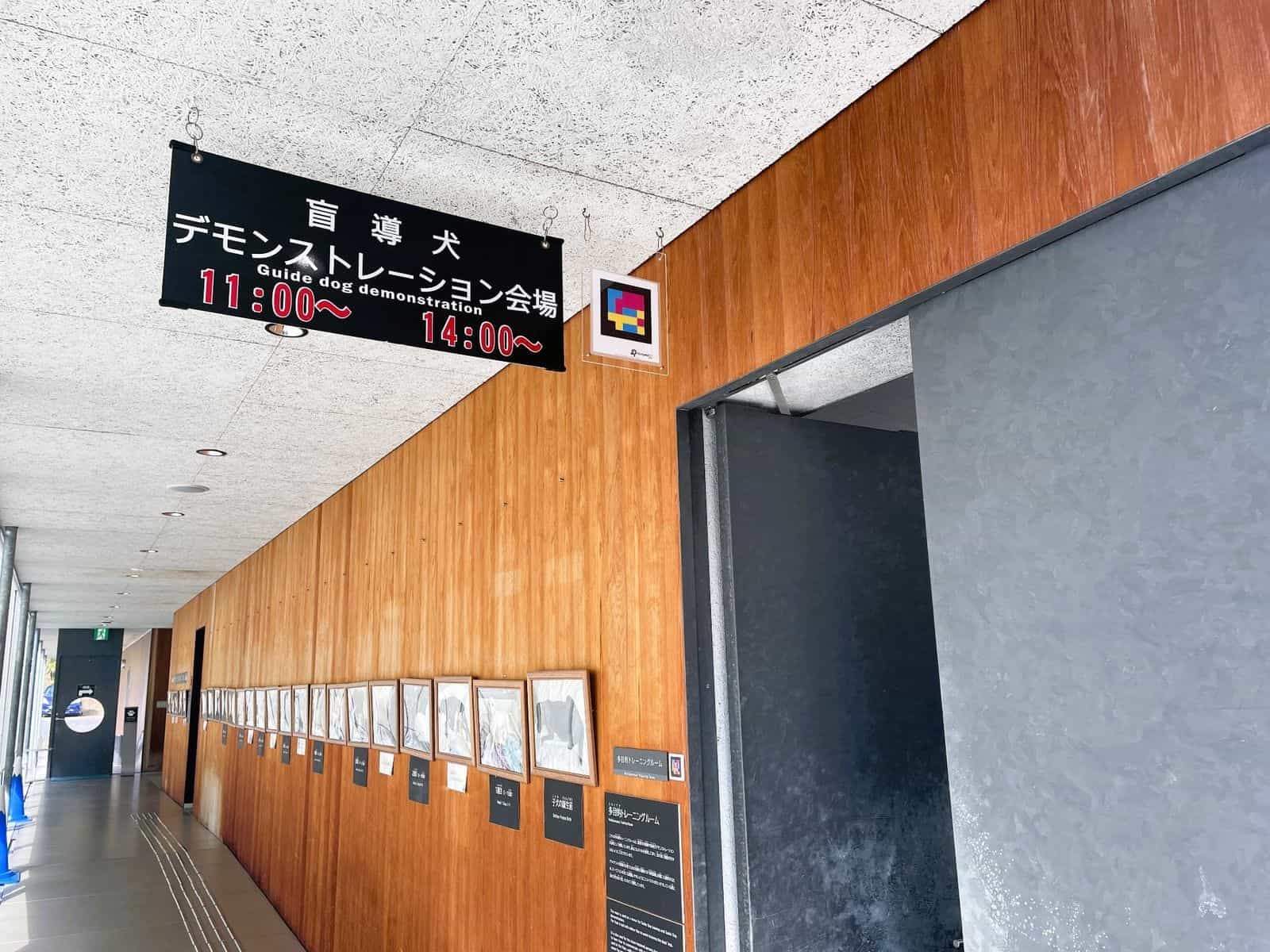 Entrance to the Multipurpose Training Room at Fuji Harness: wooden wall with framed puppy photos, black banner “盲導犬デモンストレーション会場 — Guide dog demonstration 11:00~ 14:00~” and, next to the lintel, a framed NaviLens GO code scannable from a distance
