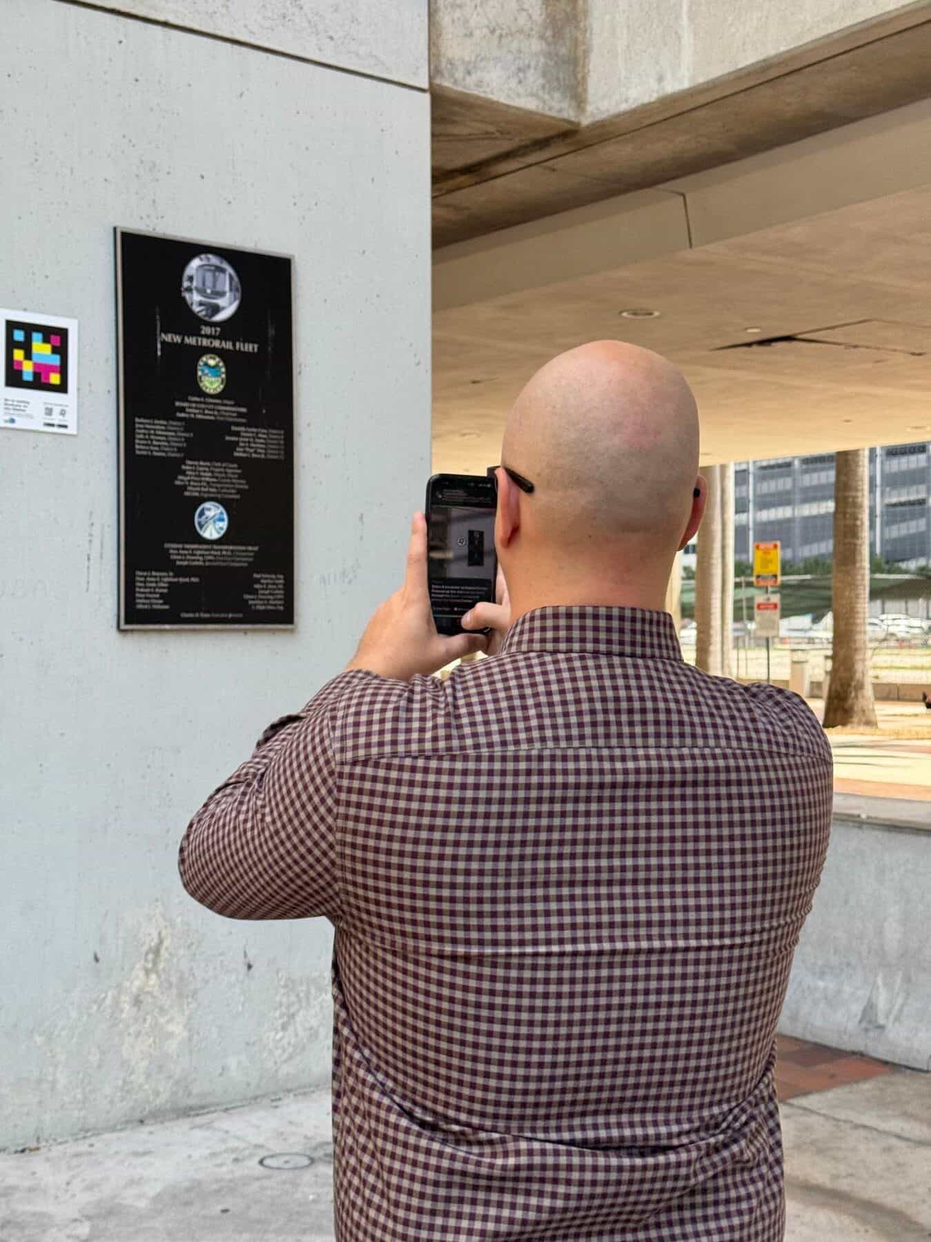 Uomo di spalle con camicia a quadri bordeaux che inquadra con il suo iPhone un codice NaviLens collocato accanto alla targa commemorativa del 2017 New Metrorail Fleet su una colonna di cemento all’uscita della stazione