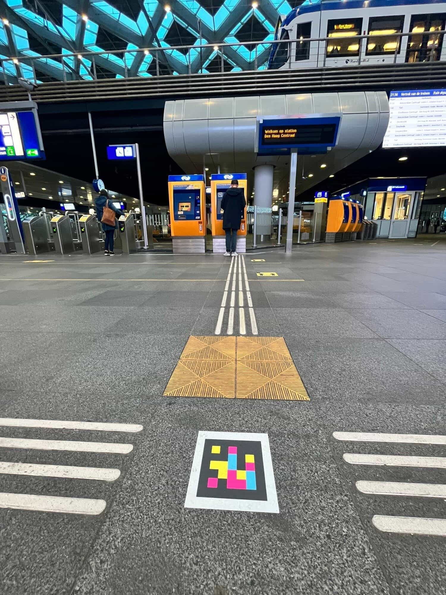 Atrio di Den Haag Centraal: linea guida tattile che termina con una piastrella direzionale e codice NaviLens fissato al pavimento, riconosciuto dall’app da diversi metri