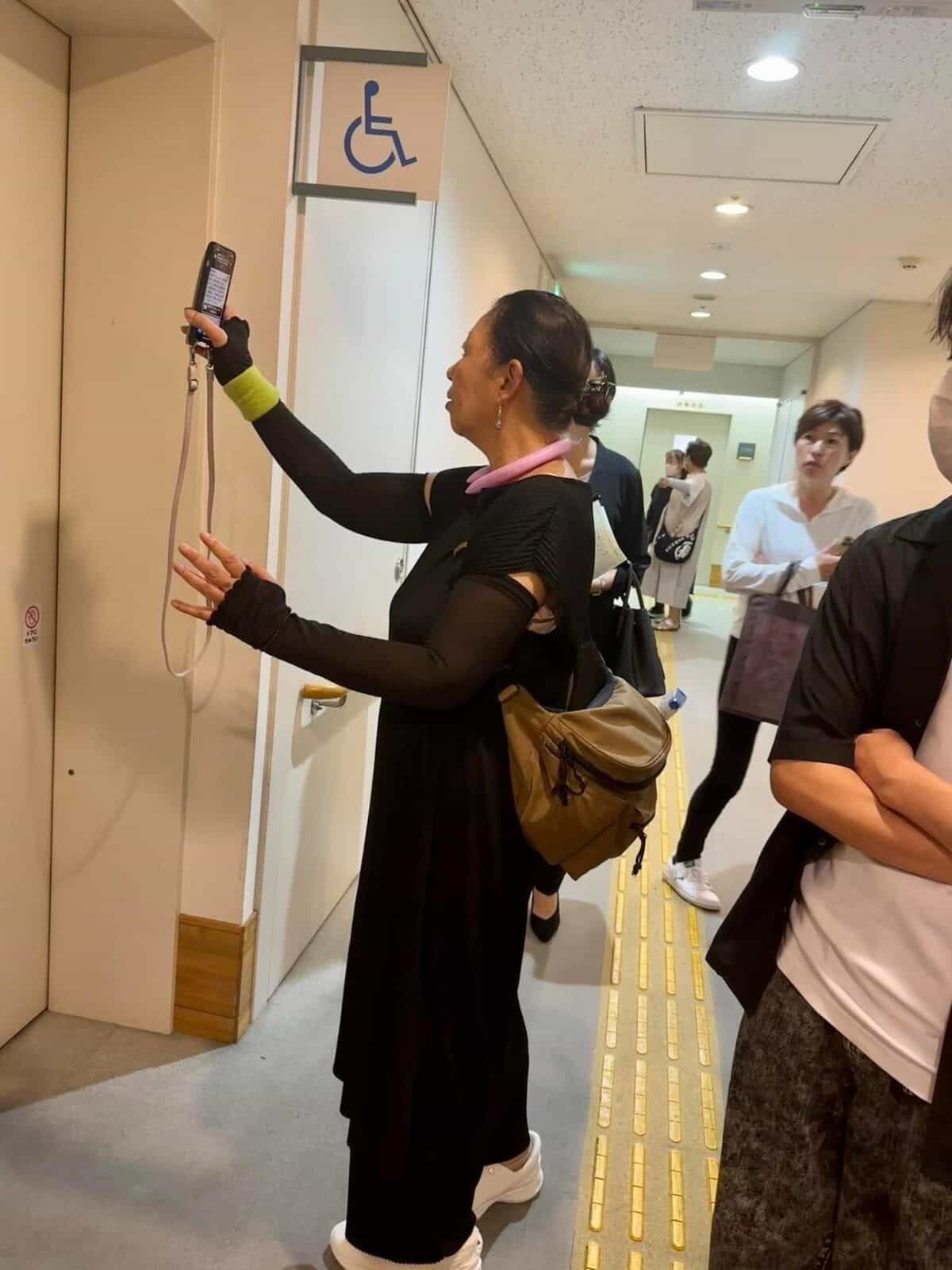 アクセシブルトイレのドア横の NaviLens 案内板にスマートフォンを向ける白杖の女性、足元には黄色い点字ブロック