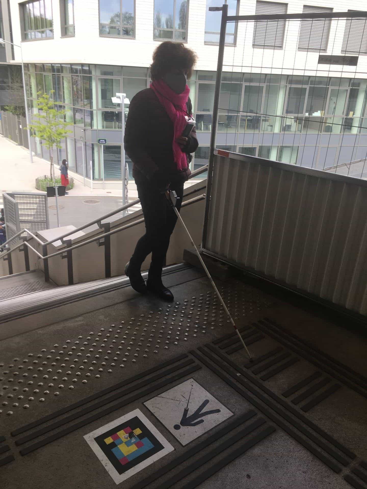 Woman with cane at the top of stairs, NaviLens code with downward arrow pictogram on the floor.