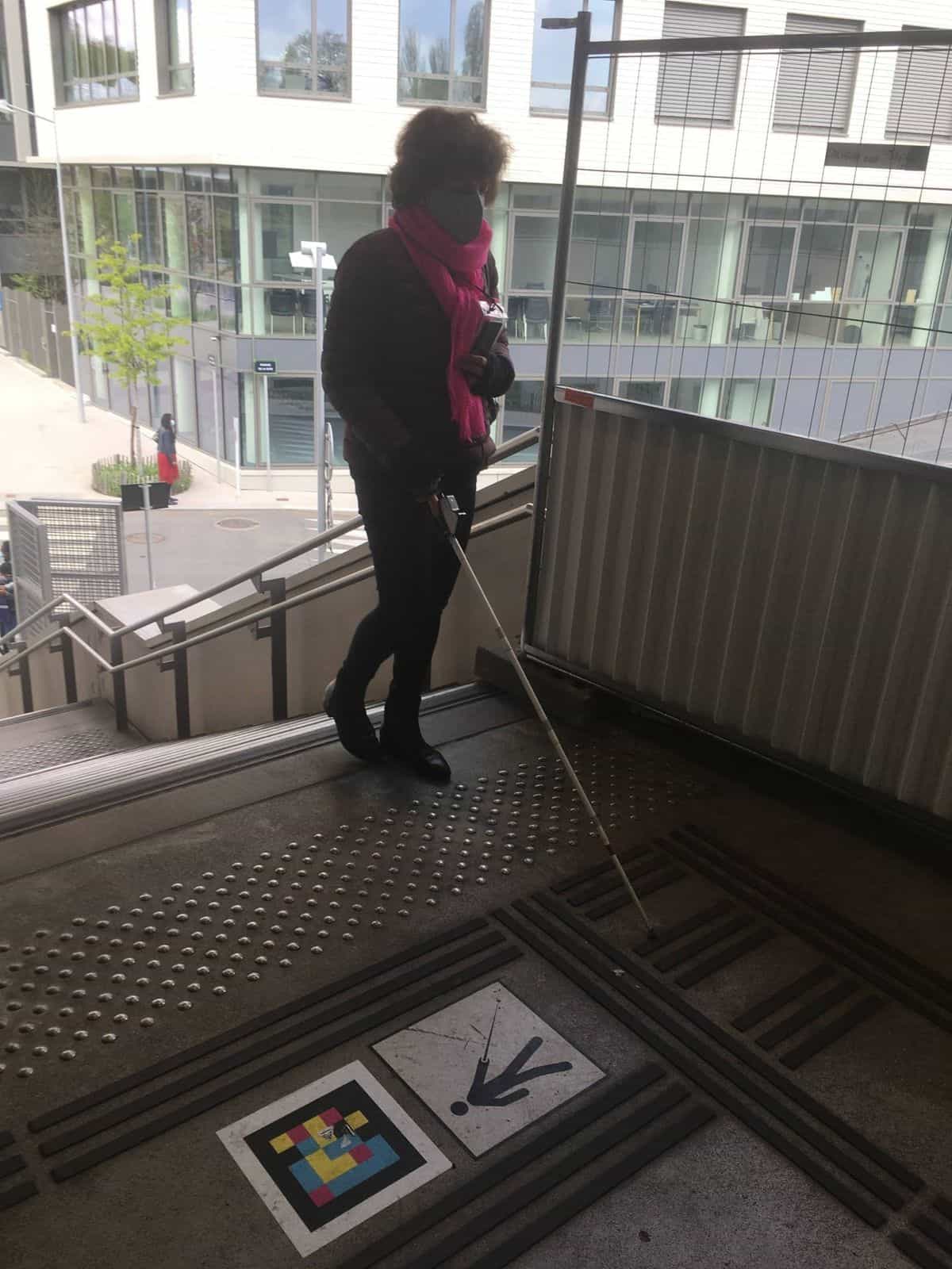 Woman with cane at the top of stairs, NaviLens code with downward arrow pictogram on the floor.