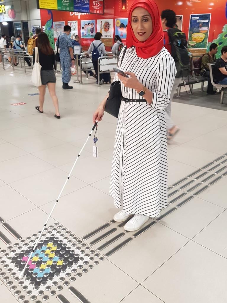 A woman in a red hijab with a white cane next to the crossing of two guide lines with a NaviLens code on the floor