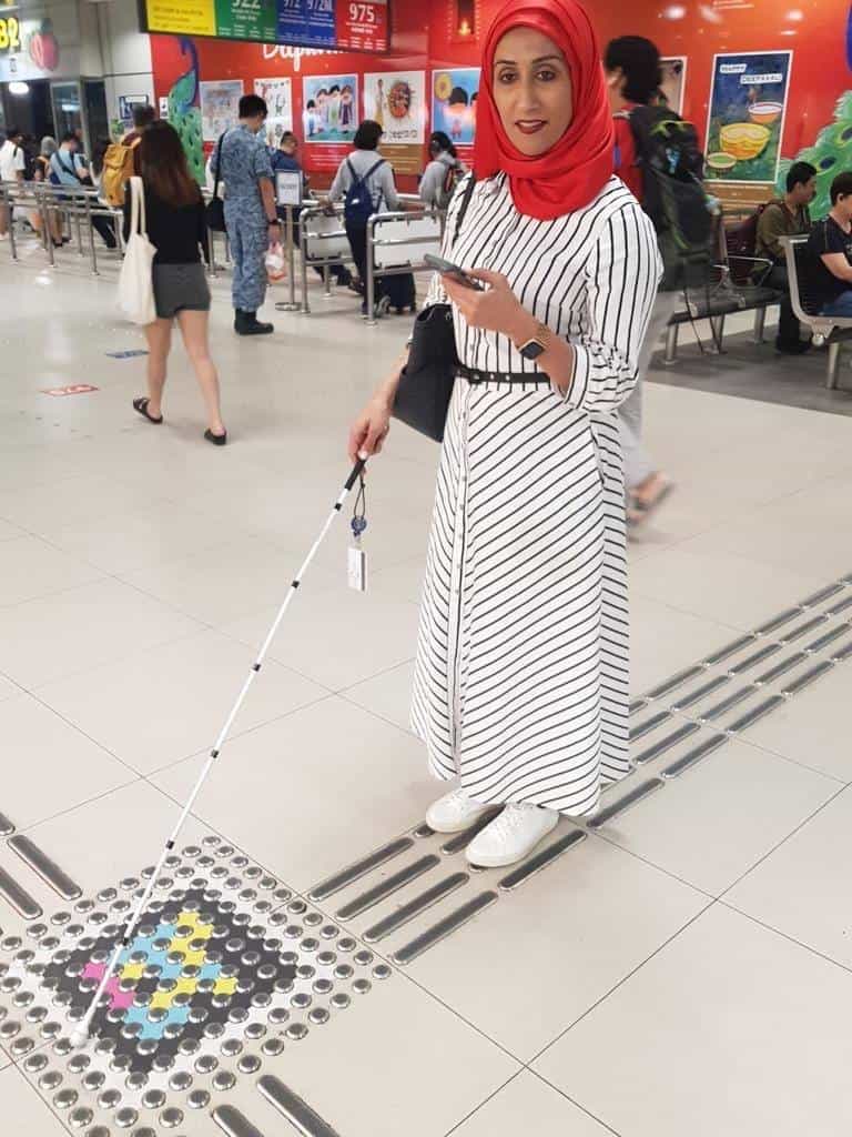 A woman in a red hijab with a white cane next to the crossing of two guide lines with a NaviLens code on the floor