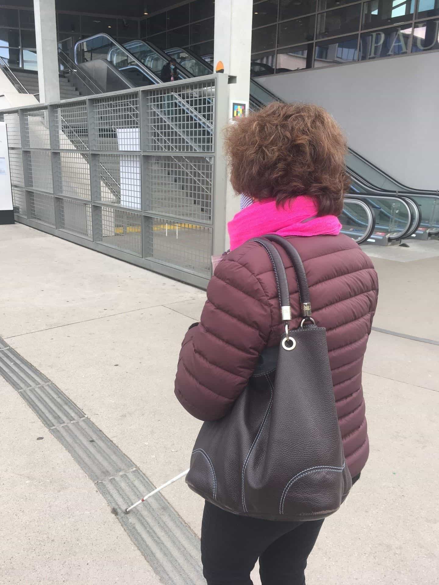 Woman with cane and backpack facing the escalators, NaviLens code on the side wall.