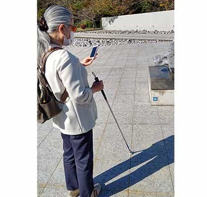 Frau mit weißem Blindenstock und dunkler Sonnenbrille schaut auf ihr iPhone im Garten des Vangi Sculpture Garden Museum, vor einer weißen Vangi-Skulptur und einem hellgrauen Granitboden