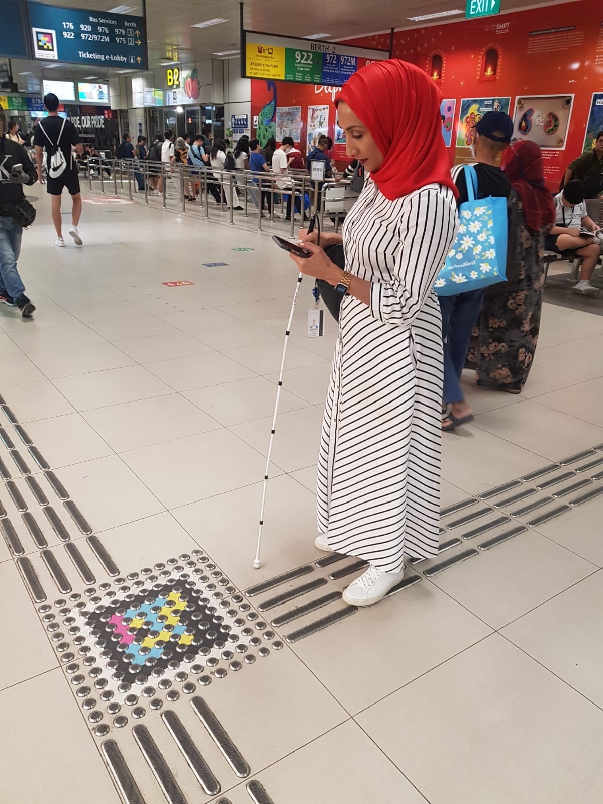 The same user scanning a large code at the crossing; in the background, the red “Berth 2” panel with services 920/922/972/975 and Deepavali posters