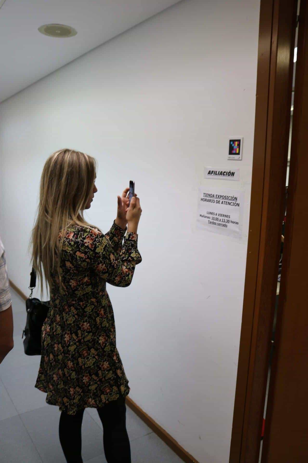 Young woman in a floral dress in a white corridor at ONCE Valladolid scanning with her phone a square colour NaviLens code placed next to the “MEMBERSHIP” and “EXHIBITION SHOP — Opening hours: Monday to Friday, mornings 10:00–13:30, afternoons closed” signs
