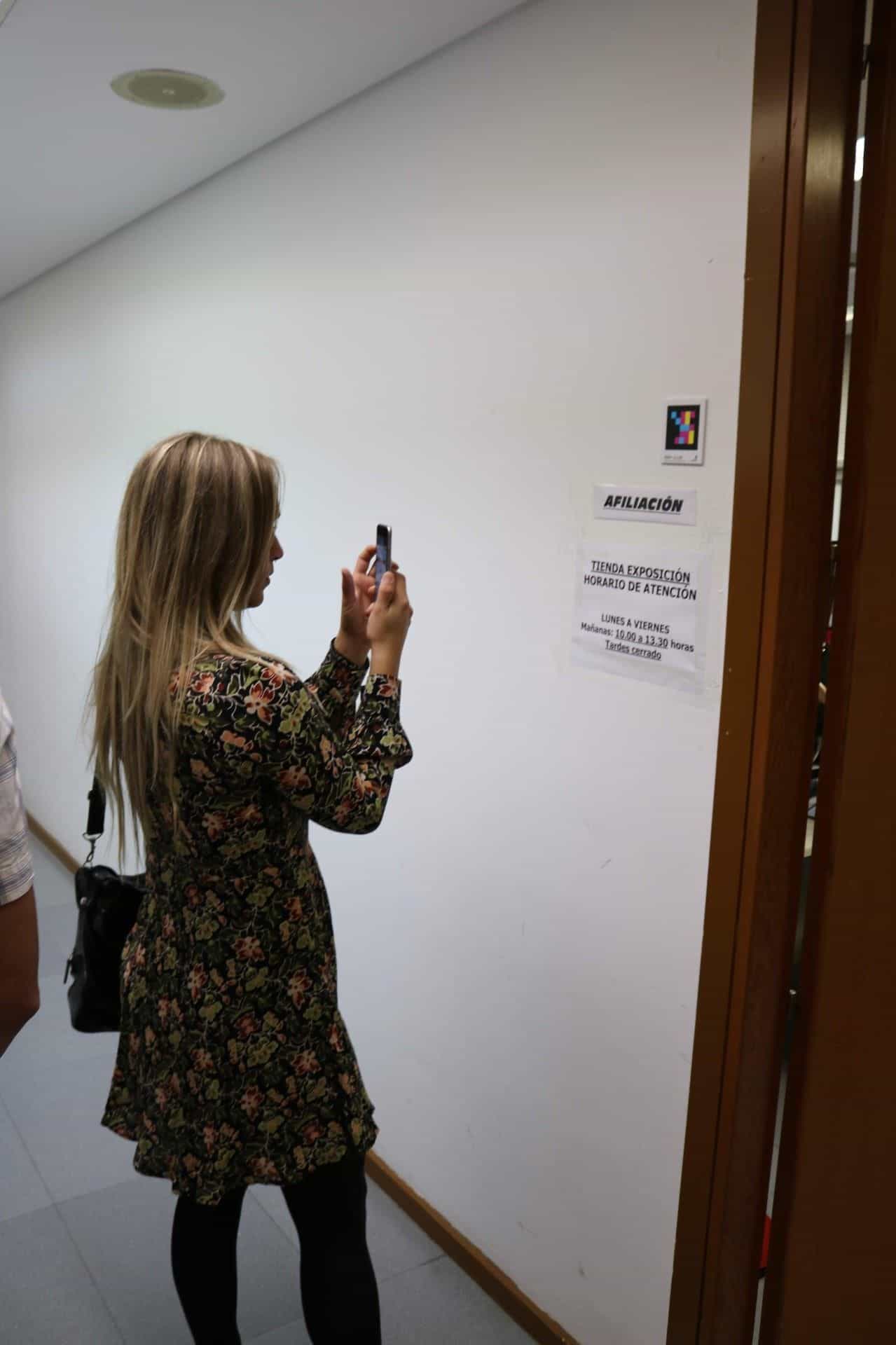 Giovane donna con vestito a fiori in un corridoio bianco dell’ONCE Valladolid che inquadra con il telefono un codice NaviLens quadrato a colori collocato accanto ai cartelli «AFILIACIÓN» e «TIENDA EXPOSICIÓN — Orario di apertura: dal lunedì al venerdì, mattina dalle 10:00 alle 13:30, pomeriggio chiuso»