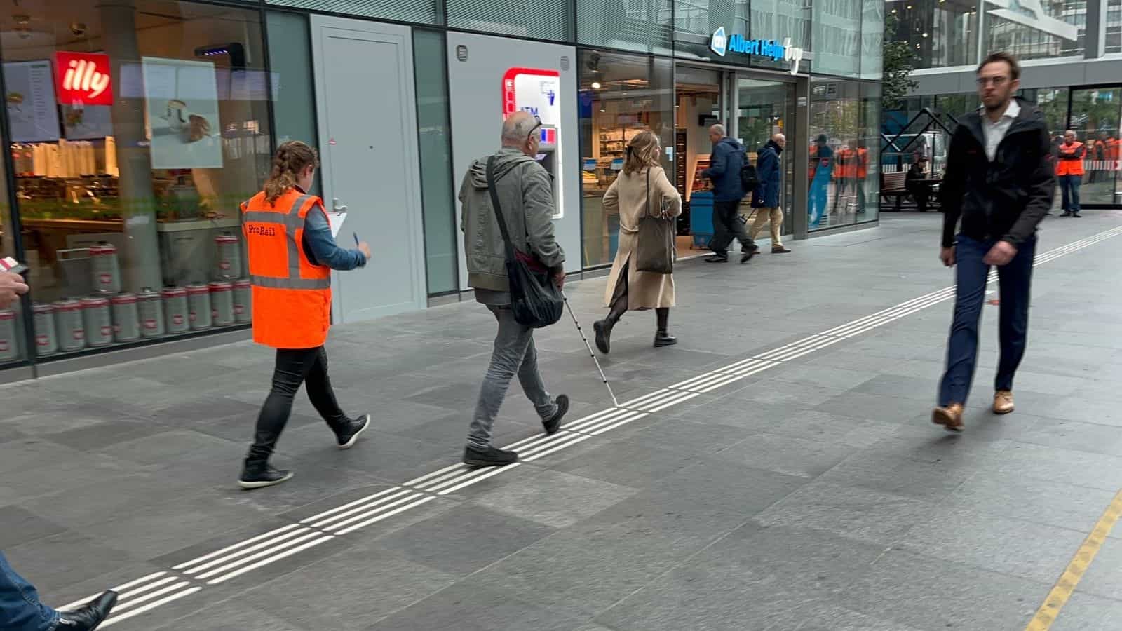 Persona con bastone bianco che percorre la linea guida tattile della galleria commerciale di Den Haag Centraal accompagnata da una volontaria con gilet arancione di ProRail che prende appunti su una tavoletta; in fondo, il supermercato Albert Heijn To Go e la caffetteria illy