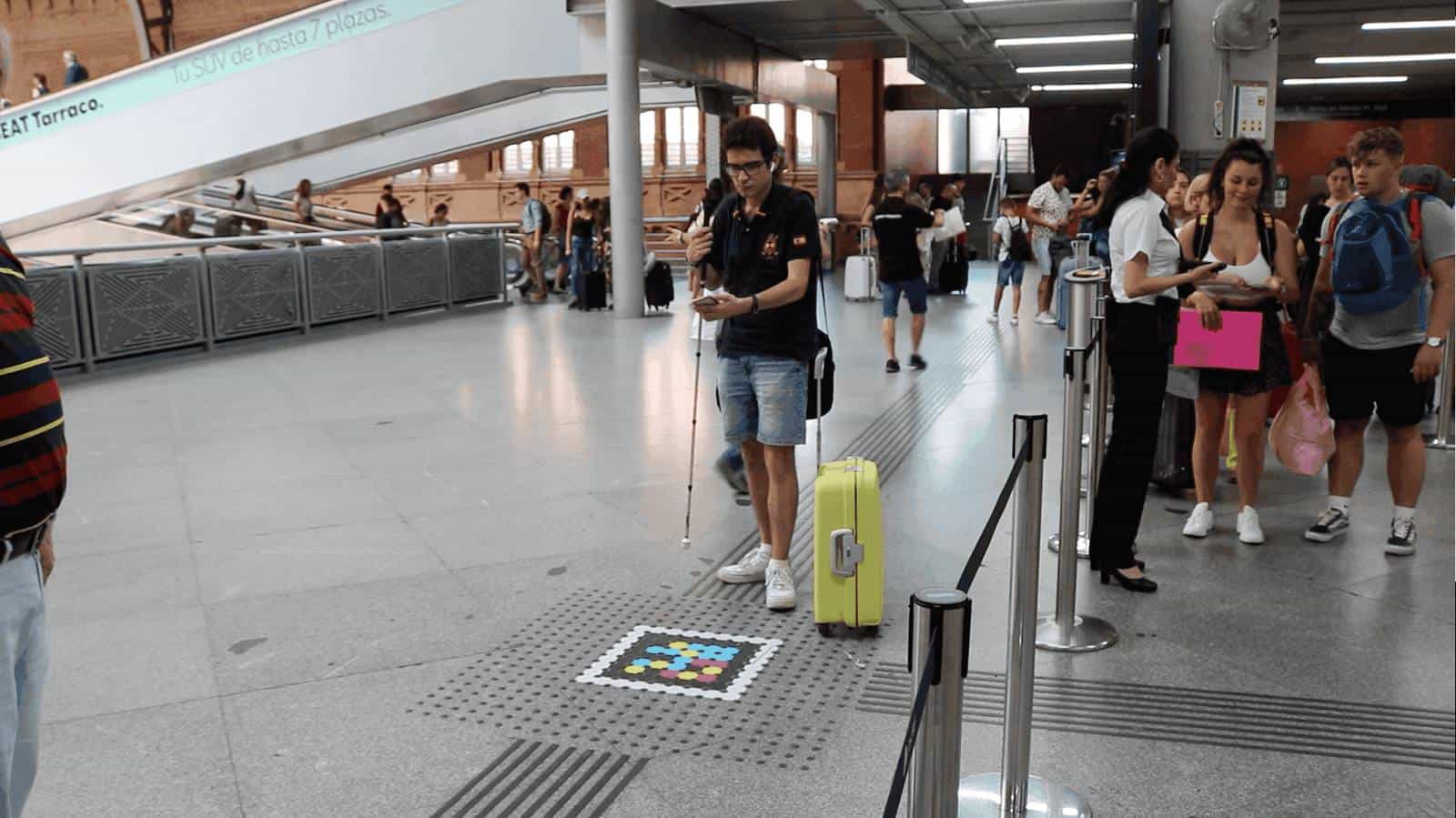User with a white cane and yellow suitcase standing on the tactile paving next to a NaviLens code on Atocha's floor; in the background, escalators, travellers and Renfe staff at the access gate.