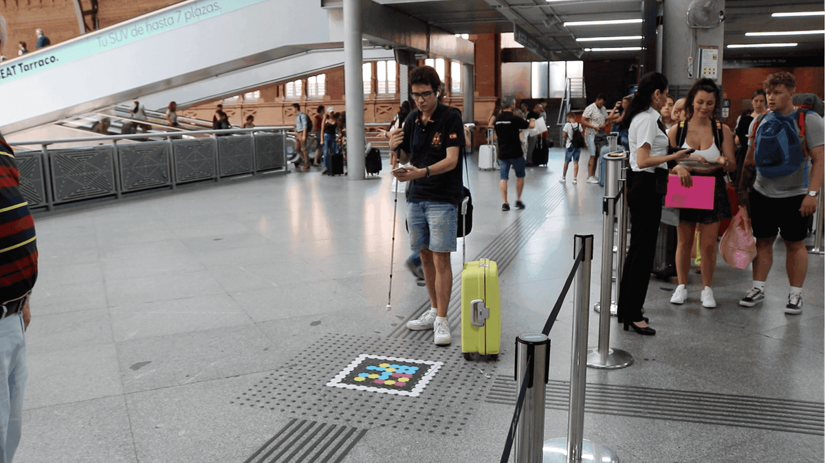 Nutzer mit weißem Stock und gelbem Koffer steht auf dem taktilen Streifen neben einem NaviLens-Code im Boden von Atocha; im Hintergrund Rolltreppen, Reisende und Renfe-Personal an der Zugangskontrolle.
