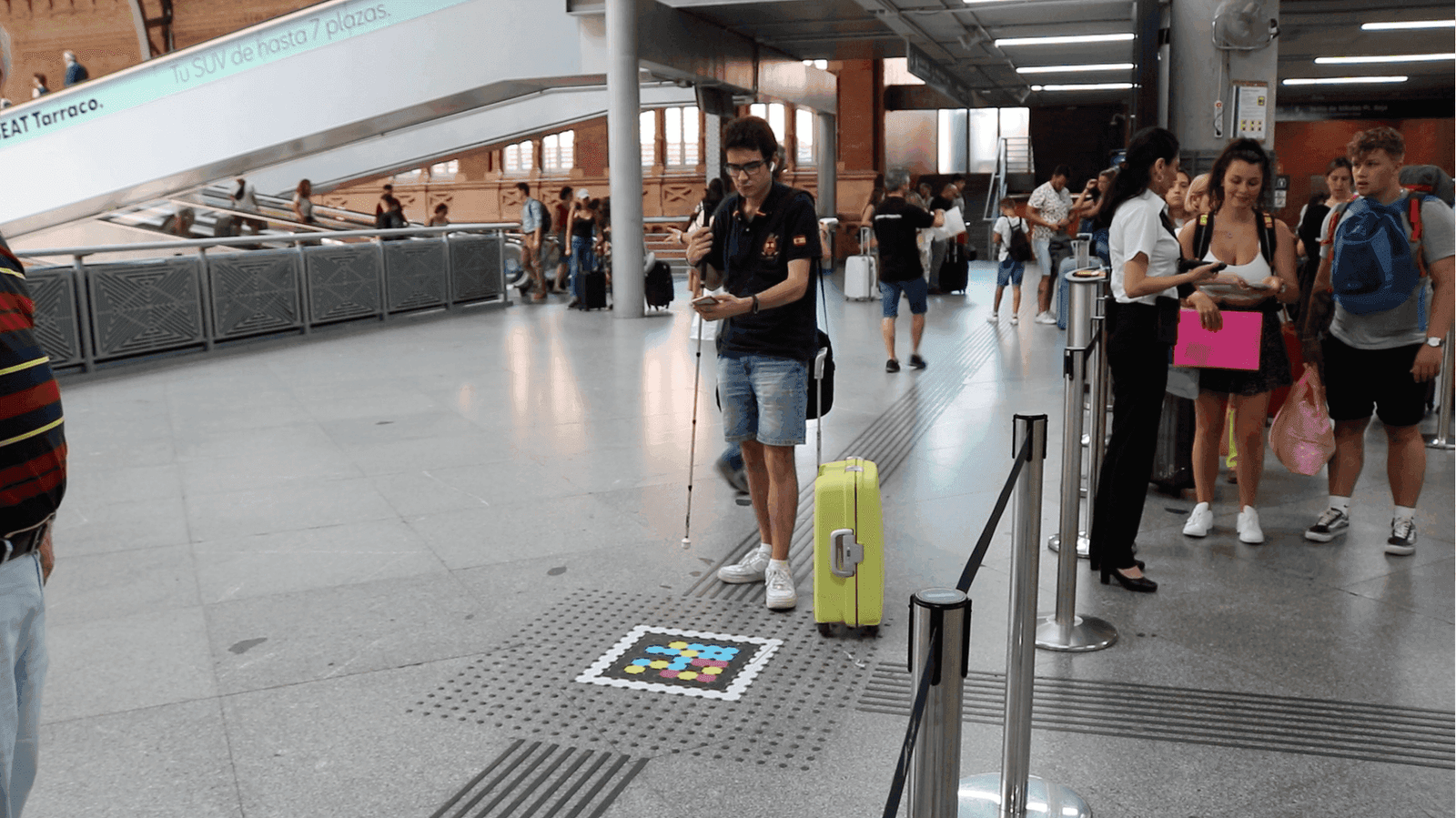 User with a white cane and yellow suitcase standing on the tactile paving next to a NaviLens code on Atocha's floor; in the background, escalators, travelers and Renfe staff at the access gate.