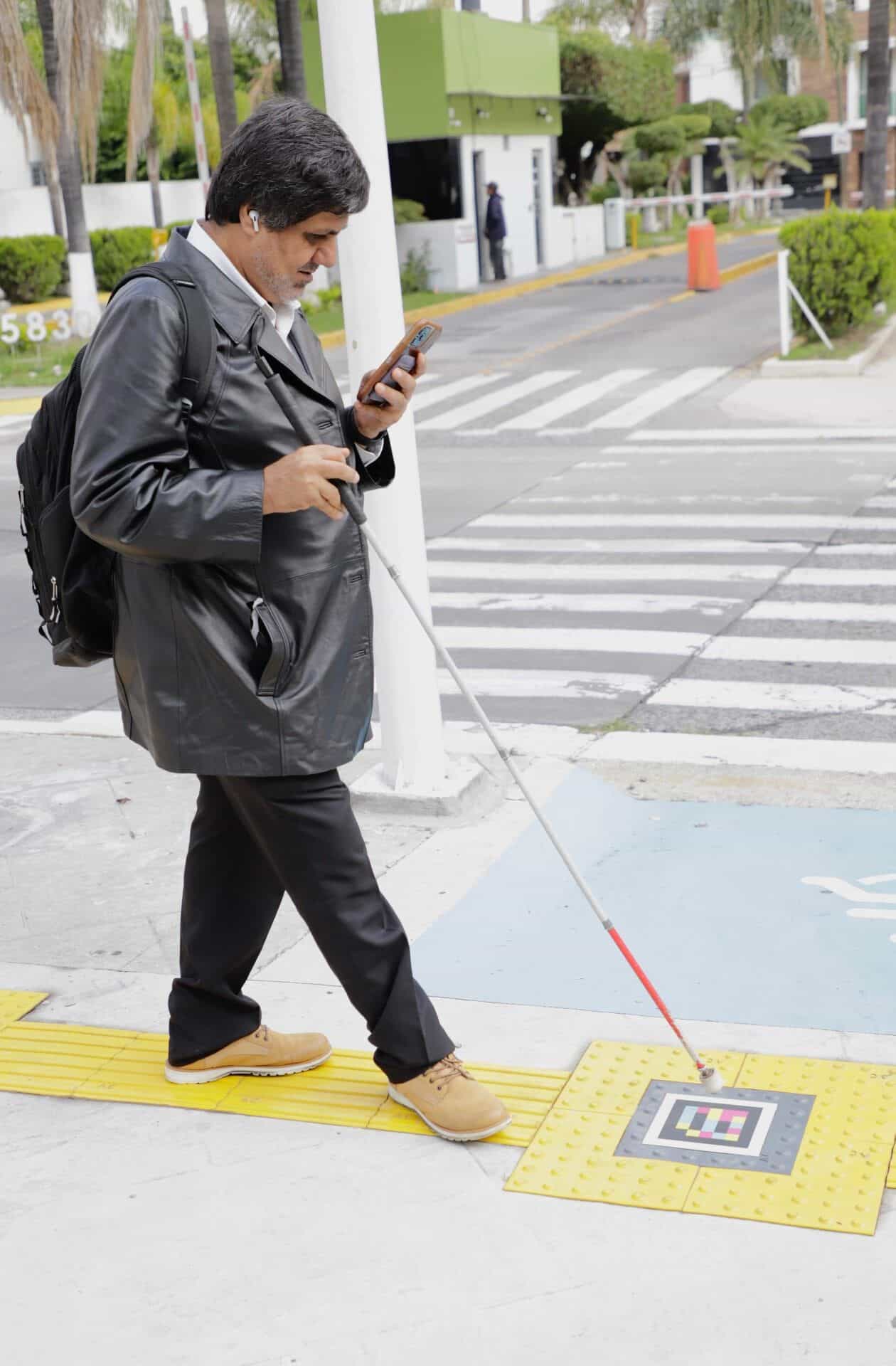 Man with a red-and-white cane and a smartphone in hand scanning a NaviLens code on a tactile tile next to the pedestrian crossing at Zapopan’s Passport Office