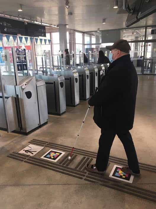 Benutzer mit weißem Stock überquert die Ticketschalter am Bahnhof Versailles-Chantiers mit NaviLens-Codes auf dem Boden